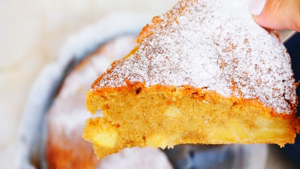A hand holding a piece of apple cake generously dusted with powdered sugar.