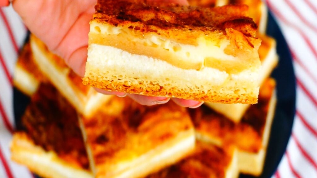 A hand holding a piece of a layered cottage cheese apple sheet cake dusted with cinnamon.